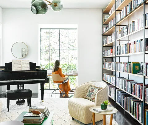 Woman in orange dress sits by a window in a bright library with bookshelves and a Yamaha piano.