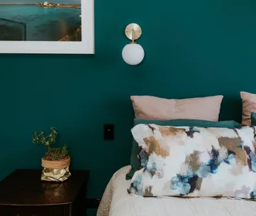 Cozy bedroom with teal wall, abstract cushion, wall lamp, framed landscape, and potted plant on bedside table.