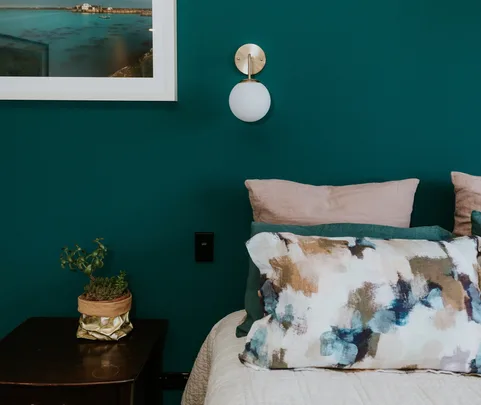 Cozy bedroom with teal wall, abstract cushion, wall lamp, framed landscape, and potted plant on bedside table.