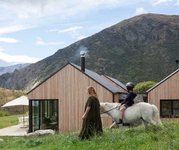 Woman and child riding a white horse near modern wooden house, with mountains in the background.