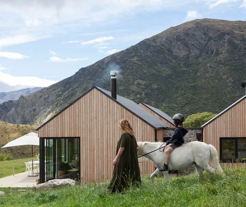 Woman and child riding a white horse near modern wooden house, with mountains in the background.