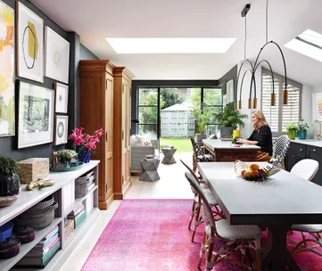 Bright modern kitchen with large windows, pink rug, and a woman seated at the table near fresh flowers and fruit.