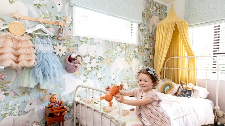 Young girl playing in a pastel unicorn-themed bedroom with toys on the bed and walls decorated with floral wallpaper.