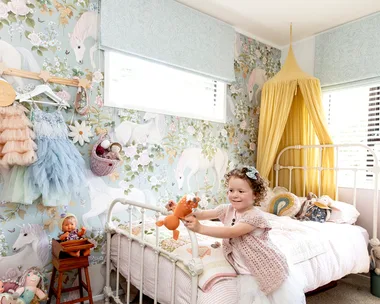 Young girl playing in a pastel unicorn-themed bedroom with toys on the bed and walls decorated with floral wallpaper.