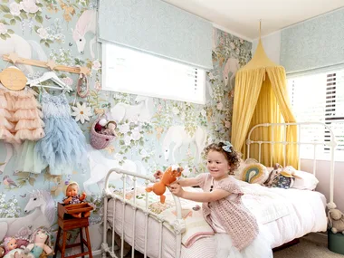 Young girl playing in a pastel unicorn-themed bedroom with toys on the bed and walls decorated with floral wallpaper.