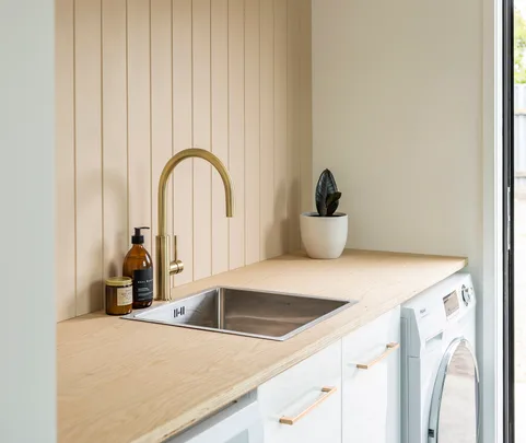 Laundry room with a sink, gold faucet, soap dispenser, plant, and white appliances under a wooden countertop.