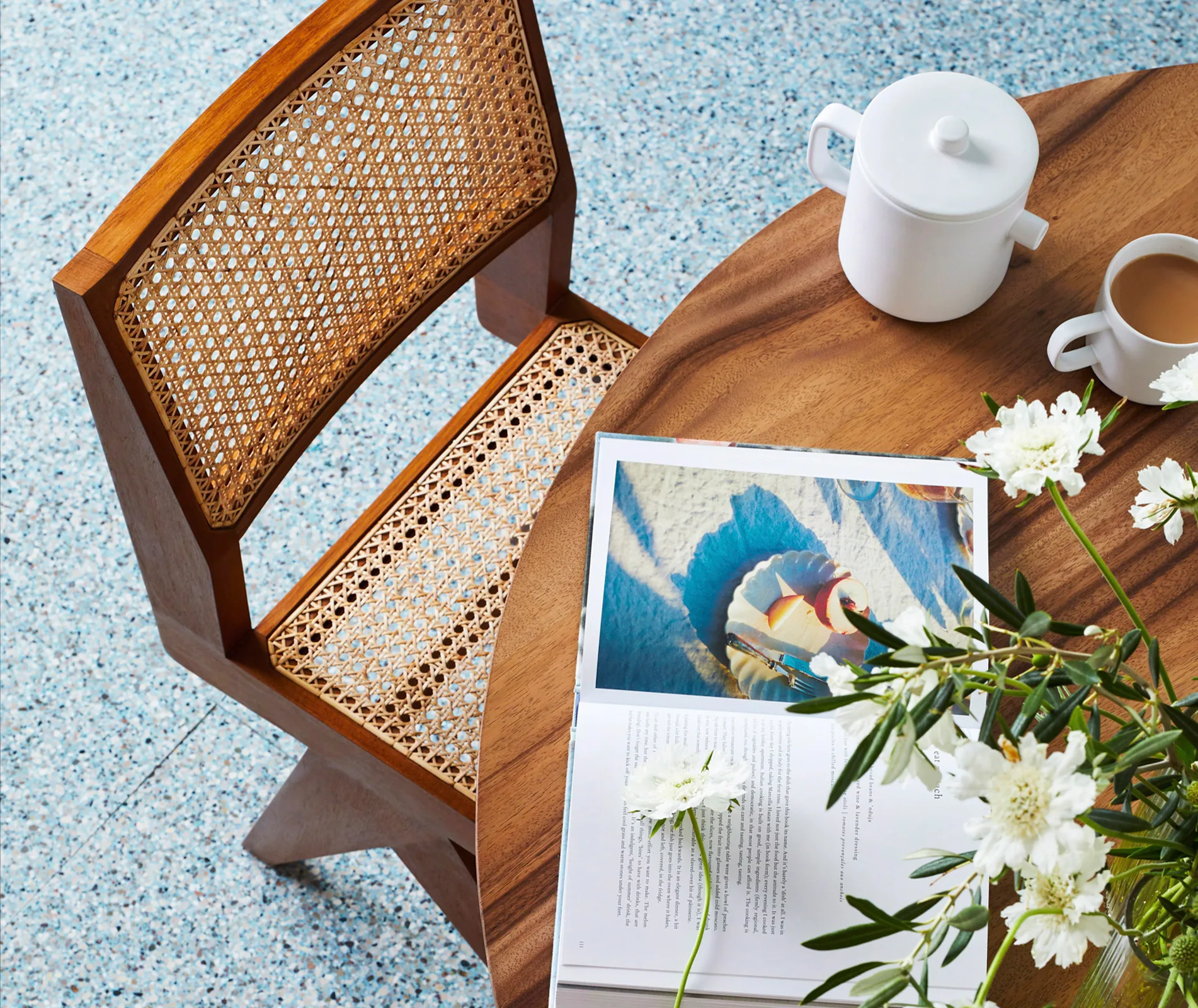 Wooden chair and table setting with a book, white flowers, a teapot, and a cup of tea.