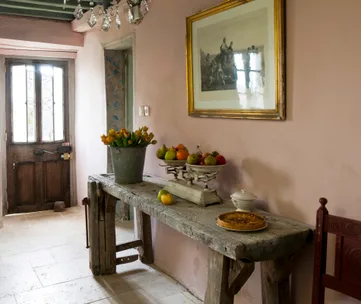 Rustic hallway with a wooden table holding flowers, fruit, and a tart; framed artwork above.