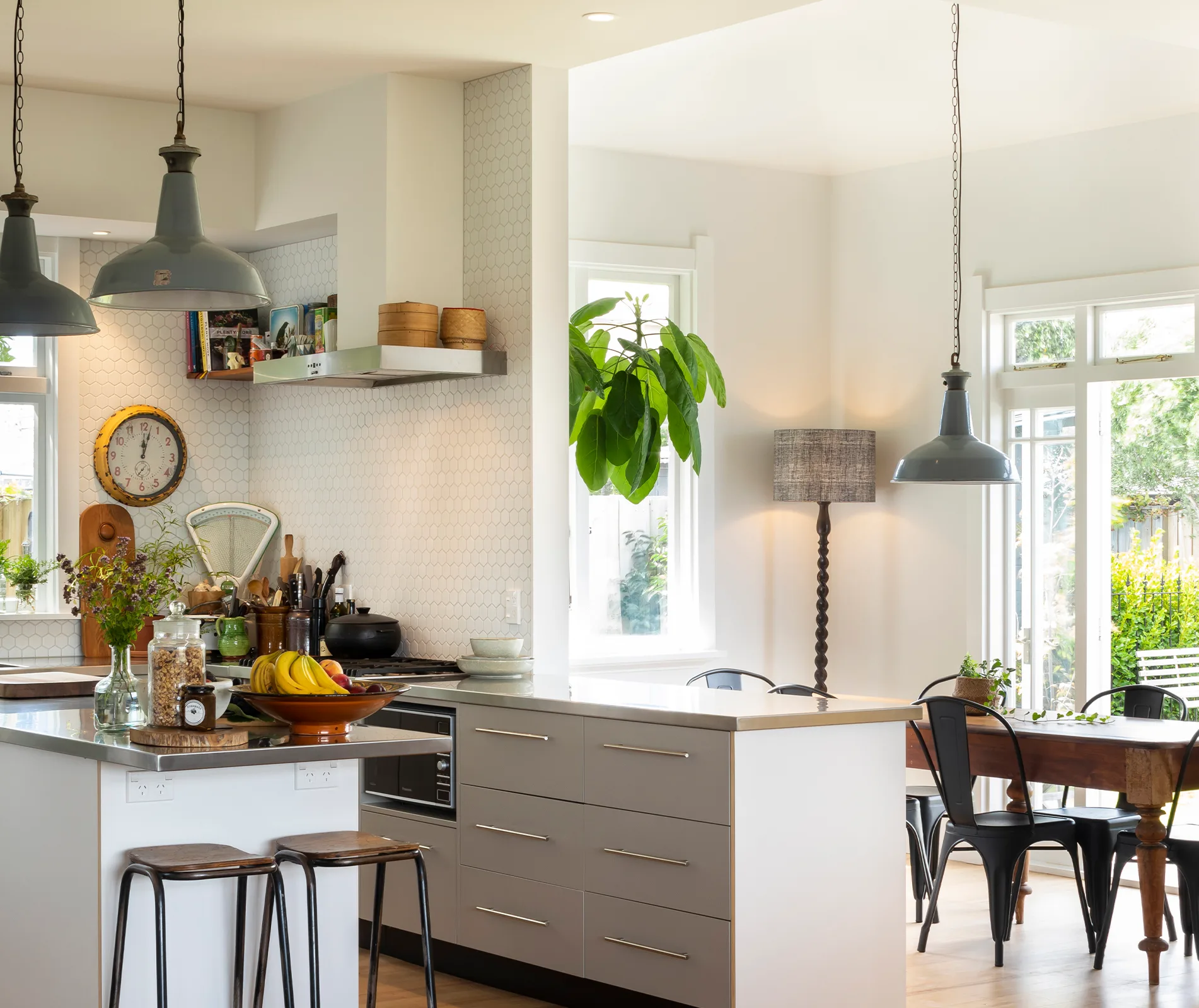 Bright kitchen with pendant lights, island with stools, plants, and dining table by large windows.