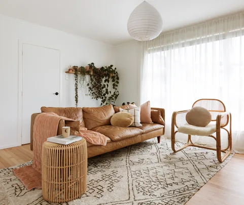 Cozy living room with a brown leather sofa, rattan chair, plants, and soft lighting.