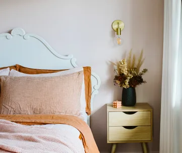 Bed with white headboard, brown bedding; wooden nightstand with vase of dried flowers, pink candle, and wall sconce above.