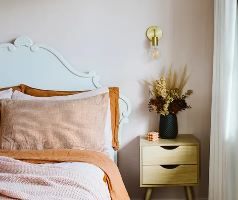 Bed with white headboard, brown bedding; wooden nightstand with vase of dried flowers, pink candle, and wall sconce above.