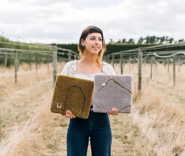 Woman in vineyard holding two textured square rugs, one brown and one grey.