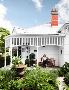 White house with porch, woman sitting on bench with dog, lush garden with potted plants in foreground.