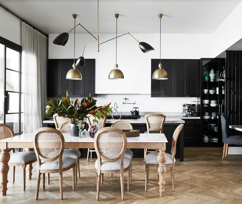 A modern dining room with a wooden table, wicker-backed chairs, black cabinets, and brass light fixtures.