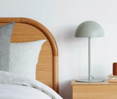 Bed with wooden headboard, plaid and striped pillows, beside a wooden nightstand with a round lamp and stacked books.