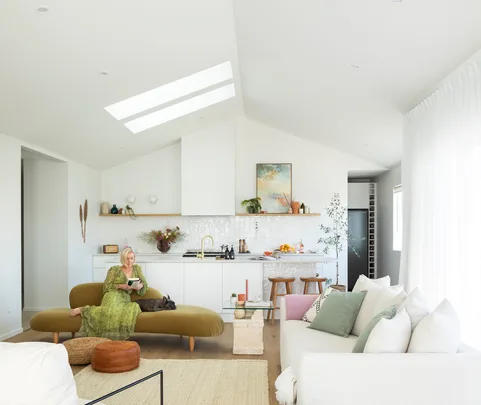 Woman in green dress sits on olive sofa with dog, reading, in bright modern living room with skylight and kitchen in background.