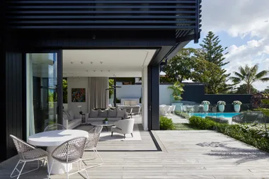 Modern house with open living room, outdoor seating, pool, and lush garden in a sunny setting.