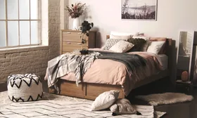Cozy bedroom with a wooden bed, pink and gray bedding, pillows, pouf, and a dresser with decor and plants.