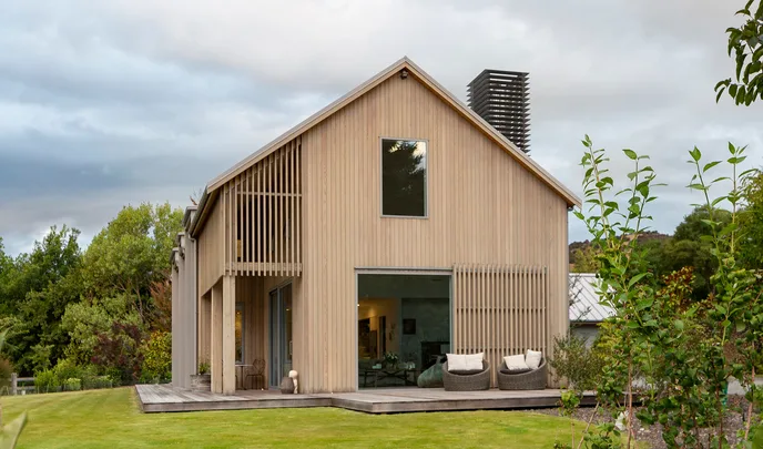 Modern wooden house with large windows, surrounded by green lawn and trees under a cloudy sky.