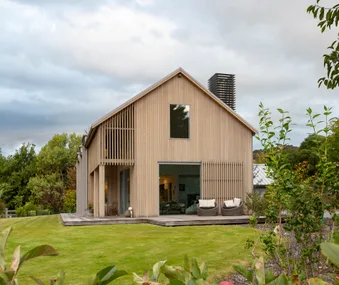 Modern wooden house with large windows, surrounded by green lawn and trees under a cloudy sky.