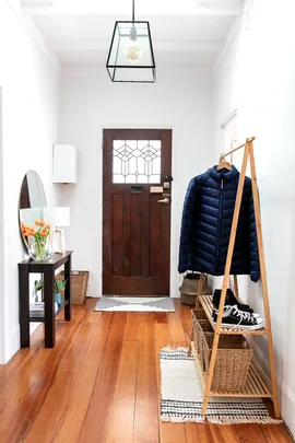 "Bright entryway with wooden floor, dark door, coat rack with jacket, sneakers, small table with lamp, and flowers."