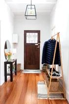 "Bright entryway with wooden floor, dark door, coat rack with jacket, sneakers, small table with lamp, and flowers."