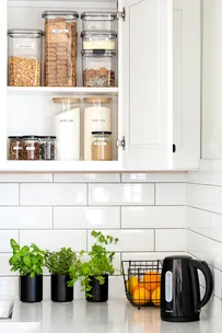 Kitchen shelves with labeled jars of cereal, sugar, and flour. Counter with herbs, oranges, and a black kettle.