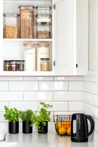 Kitchen shelves with labeled jars of cereal, sugar, and flour. Counter with herbs, oranges, and a black kettle.