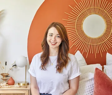 A woman with long hair smiles while sitting in a room with a sunburst mirror and modern decor.
