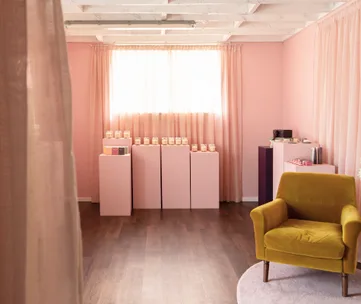Cozy room with pink walls, a yellow armchair, and products displayed on pink pedestals lit by soft natural light.