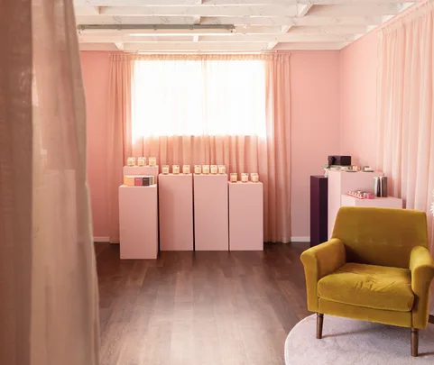 Cozy room with pink walls, a yellow armchair, and products displayed on pink pedestals lit by soft natural light.
