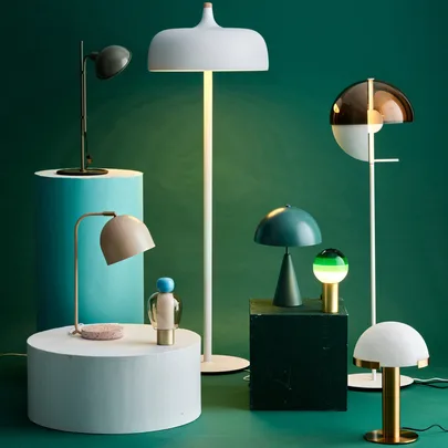 Modern table and floor lamps in various designs and colors on pedestals, displayed against a teal background.