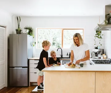 A woman and two children in a modern kitchen prepare food on the counter. Bright and airy setting with plants and appliances.