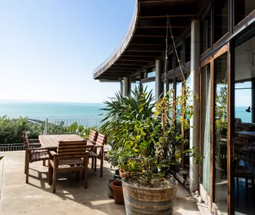 Patio with wooden furniture, potted plants, and ocean view under clear blue sky.