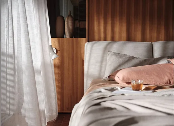 Cozy bedroom with pale pink and beige pillows on a bed, wooden headboard, white curtains, and soft natural light.