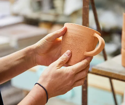 Hands holding a handcrafted, unglazed clay mug in a workshop.