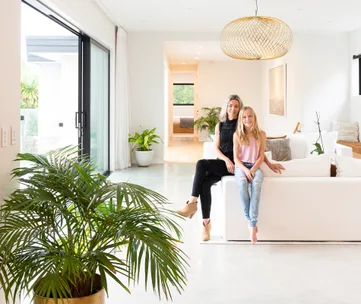 Two people sit on a white sofa in a modern, bright living room with plants and large windows.