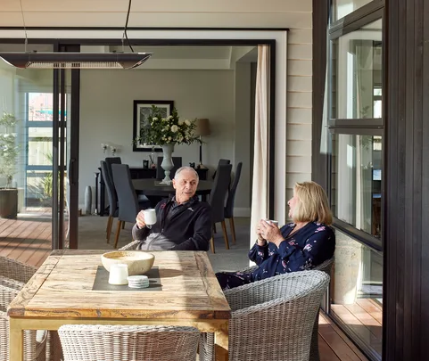 Elderly couple sitting at a patio table, having coffee and chatting outside a modern home.