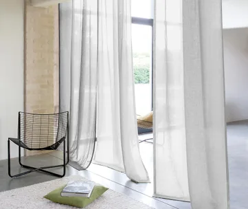 White curtains hang by a large window, with a black wire chair and a green cushion on a light rug.