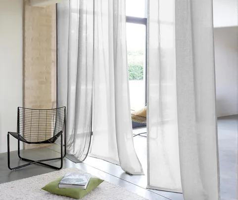 White curtains hang by a large window, with a black wire chair and a green cushion on a light rug.