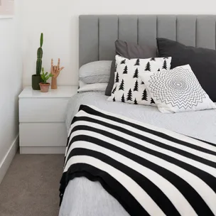 Modern bedroom with a grey headboard, black and white pillows, striped throw, and potted plants on a bedside table.