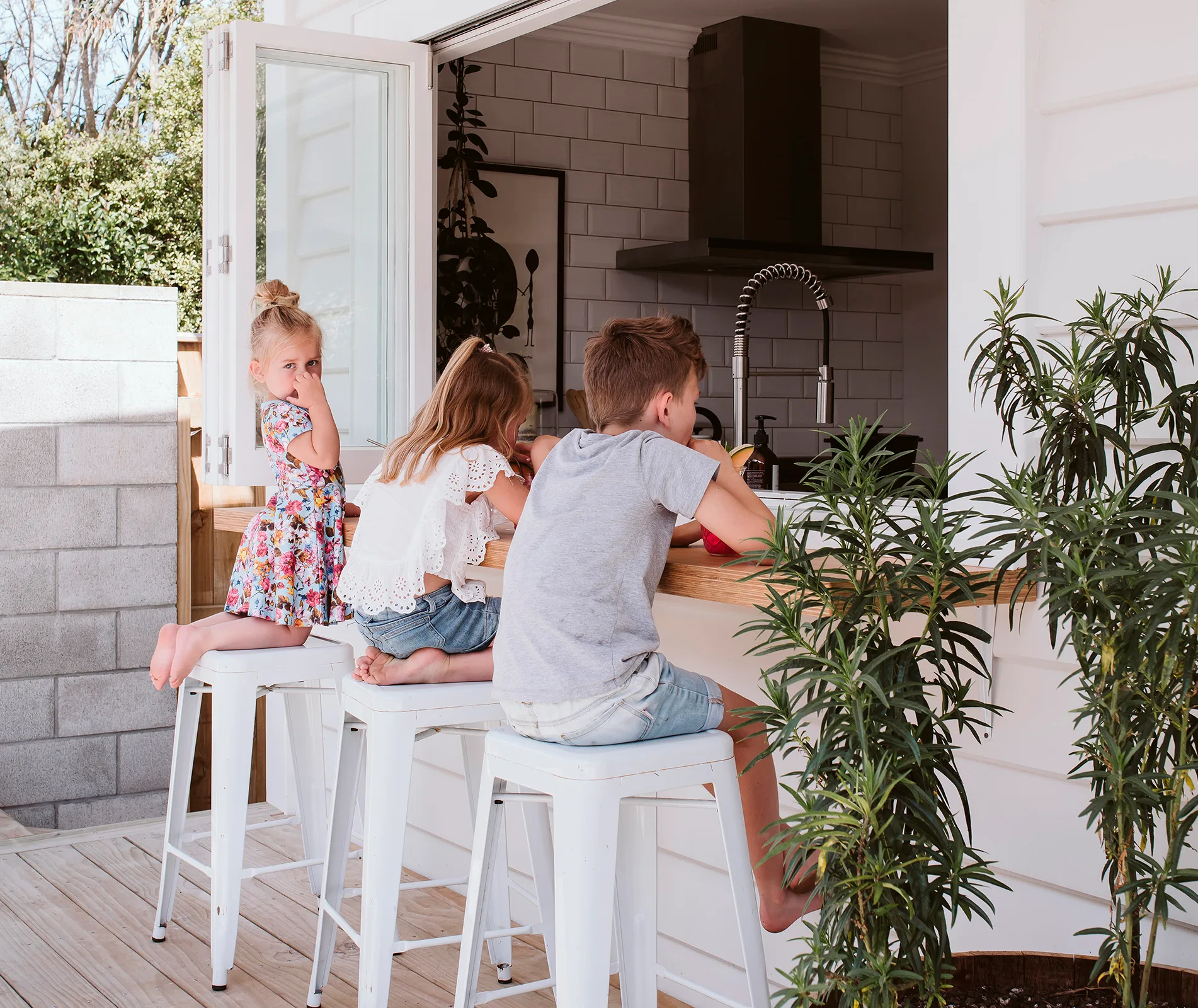 Three children sitting on stools at an outdoor kitchen counter, one girl looking at the camera, surrounded by greenery.