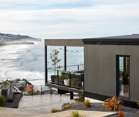 Cliffside modern house with ocean views, featuring a glass balcony and potted plants under overcast skies.