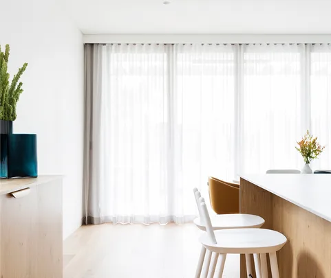 Modern minimalist dining area with light wood furniture, white chairs, a vase with flowers, and large sheer curtains.