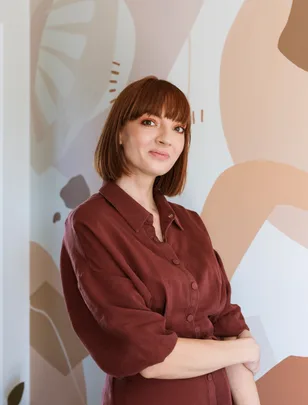 Person with red bob hairstyle in maroon outfit, standing in front of abstract beige and gray wall art.