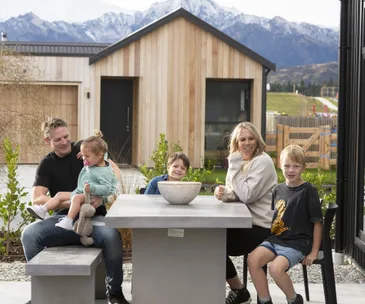 A family sits around an outdoor table in front of a modern wooden house with mountains in the background.