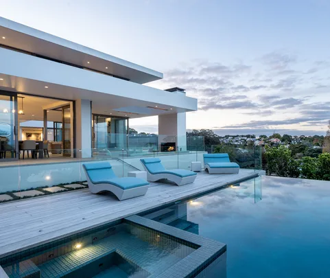 Modern house with glass walls, blue lounge chairs, and an infinity pool overlooking a scenic landscape at sunset.