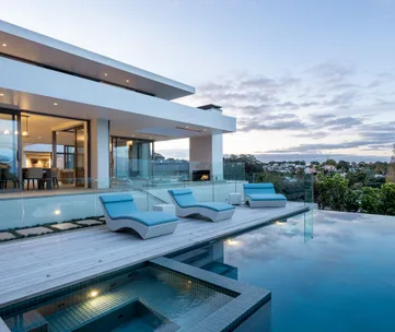 Modern house with glass walls, blue lounge chairs, and an infinity pool overlooking a scenic landscape at sunset.