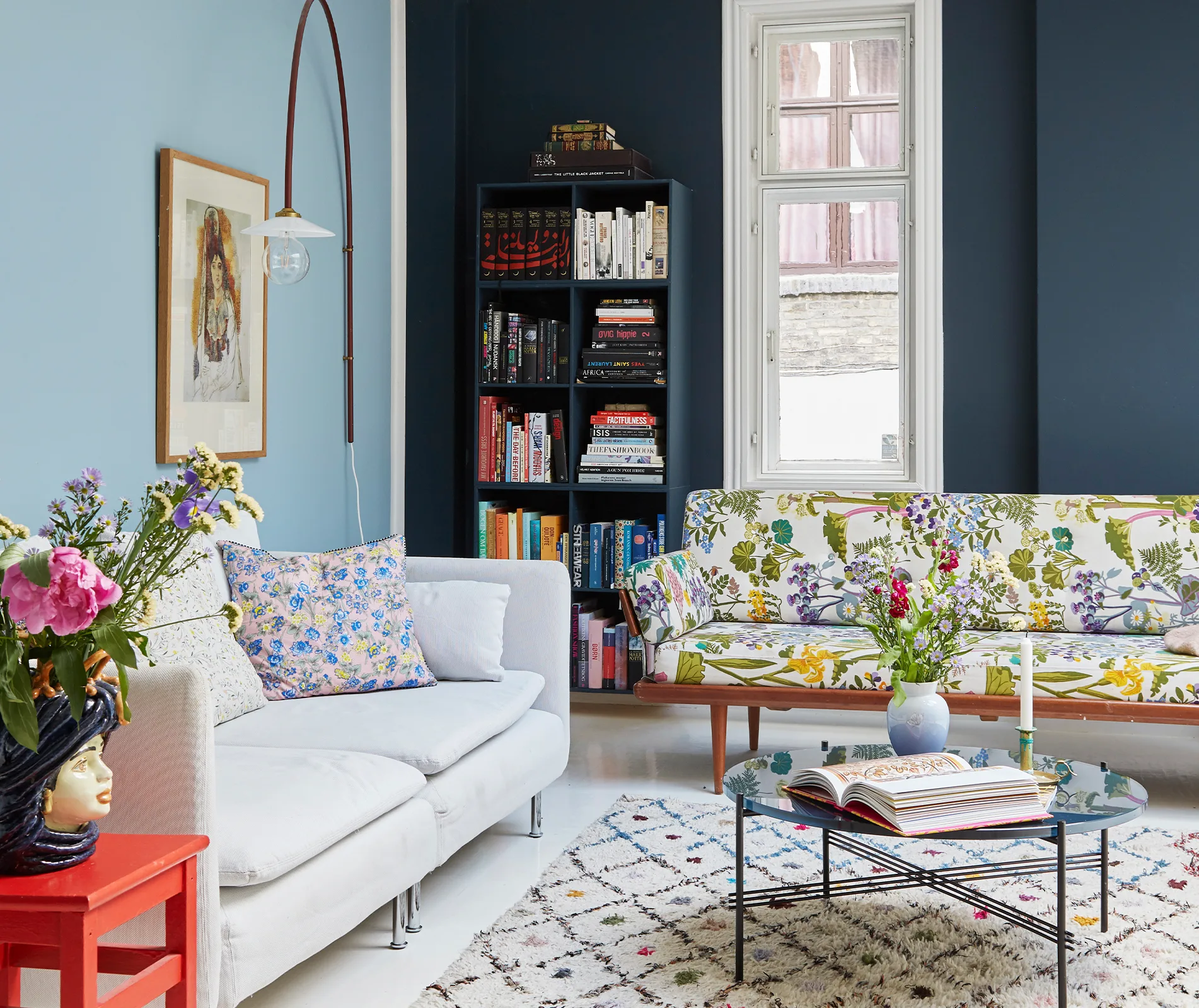 A cozy living room with floral cushions, bookshelves, a painting, and a glass coffee table with a vase of flowers.